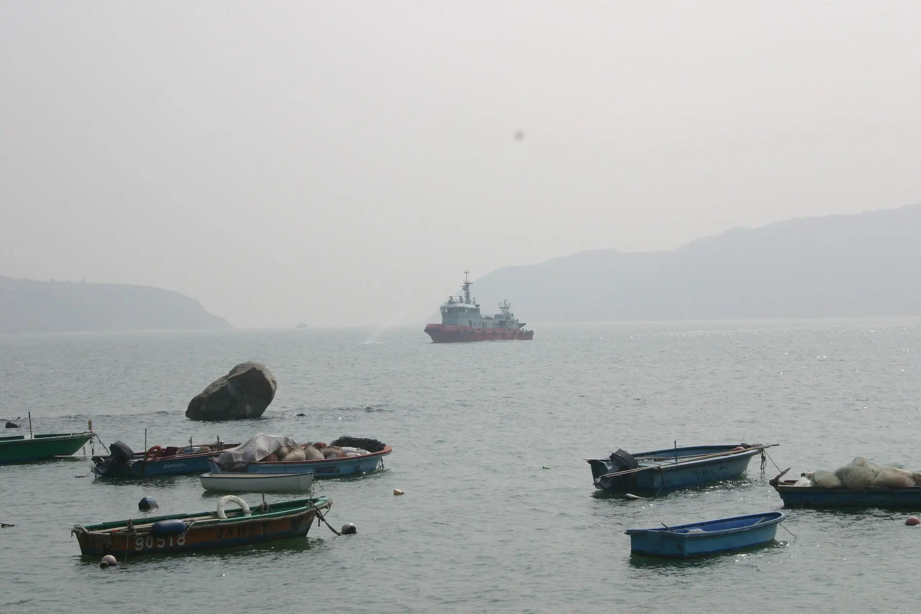 Red ship spraying water beyond small moored fishing boats on a hazy sea
