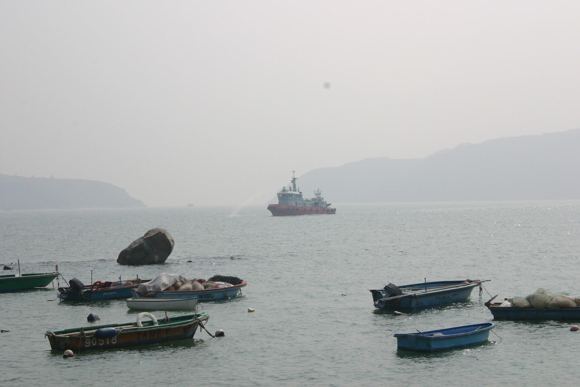 Red ship spraying water beyond small moored fishing boats on a hazy sea