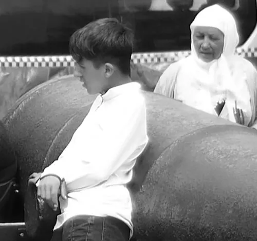 Crop of the leaning boy in a white shirt and the older woman in a white headscarf, both looking down